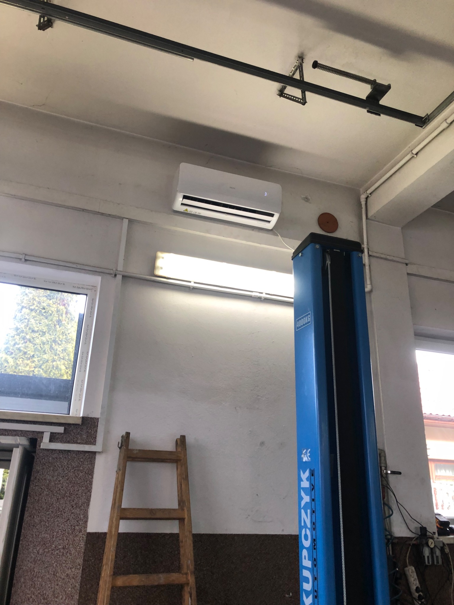 Wnętrze warsztatu z zamontowanym klimatyzatorem na ścianie, widoczny podnośnik samochodowy, drabina i okno.