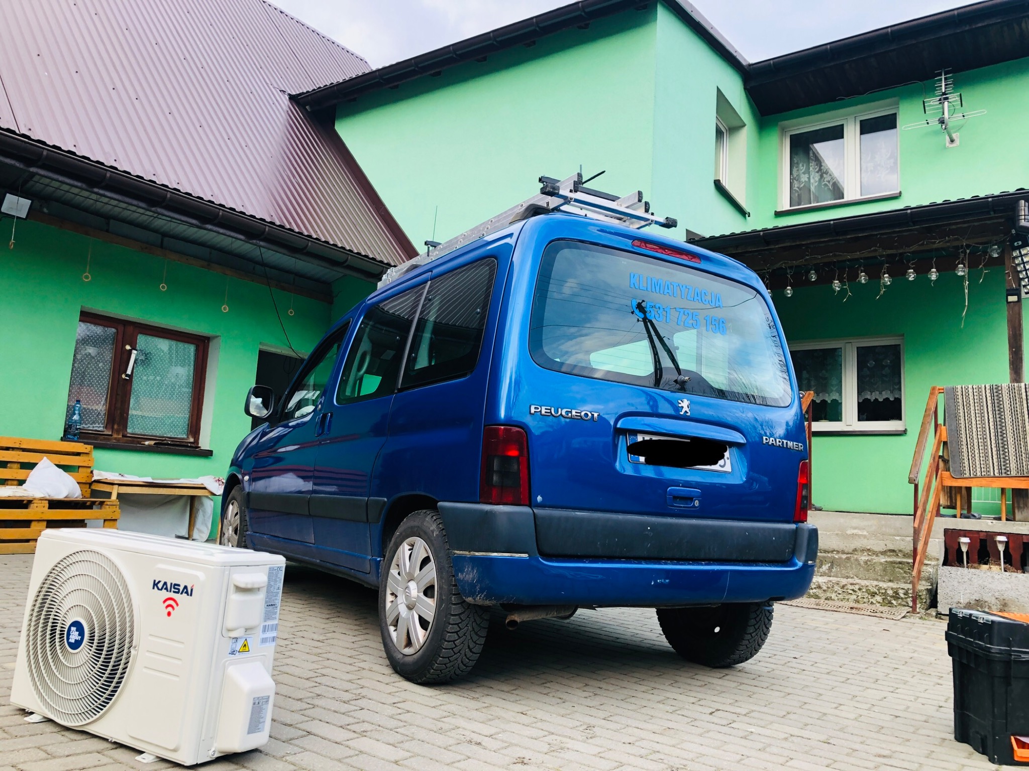 Jednostka zewnętrzna klimatyzacji Kaisai leżąca na kostce brukowej przed niebieskim Peugeot Partner z drabiną na dachu, na tle zielonego domu z brązowym dachem.