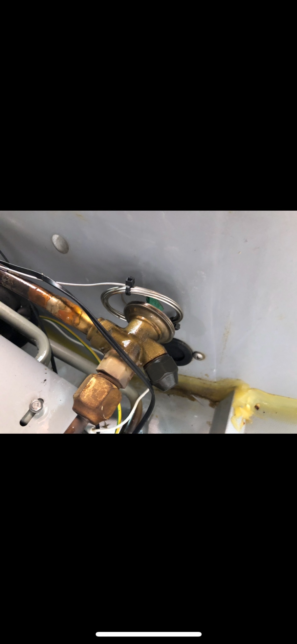 Mosiężny zawór rozprężny klimatyzacji z zielonym elementem, przymocowany opaską zaciskową, widoczne miedziane rurki i okablowanie, brudne tło.