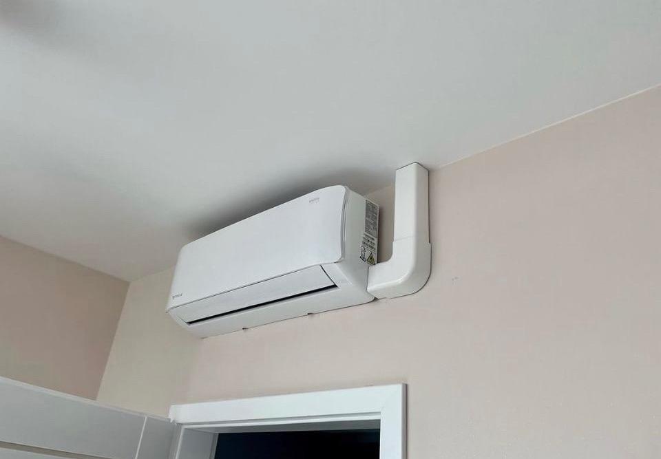 Biały klimatyzator ścienny zamontowany nad framugą drzwi, z widoczną instalacją odprowadzającą skropliny w białej obudowie.