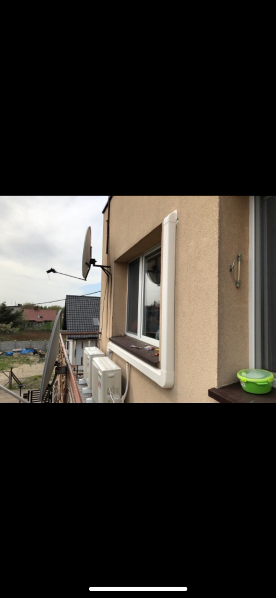 Dwa białe klimatyzatory zamontowane na balkonie budynku mieszkalnego, widoczne z zewnątrz, z anteną satelitarną i zielonym pojemnikiem na parapecie okiennym.