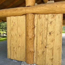 Drewniana konstrukcja altany z bali, widoczne połączenia bali i desek, podstawa kolumny z kamieni.