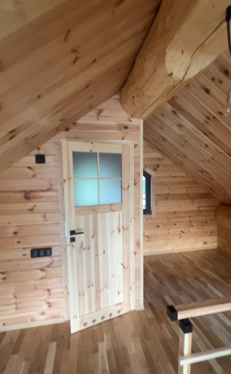 Wnętrze poddasza wykończonego drewnem: ściany z bali, drewniana podłoga i drzwi z przeszkleniami, widoczne gniazdka elektryczne, małe okno oraz fragment konstrukcji dachu z grubą belką.