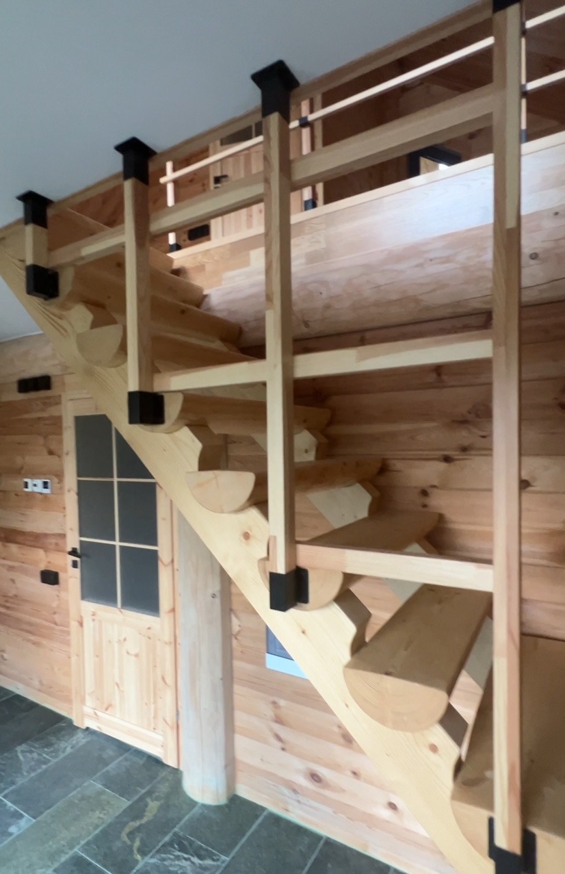 Wnętrze domu z bali: drewniane schody z czarnymi akcentami, prowadzące na antresolę, widoczne drzwi wejściowe i kamienna podłoga.