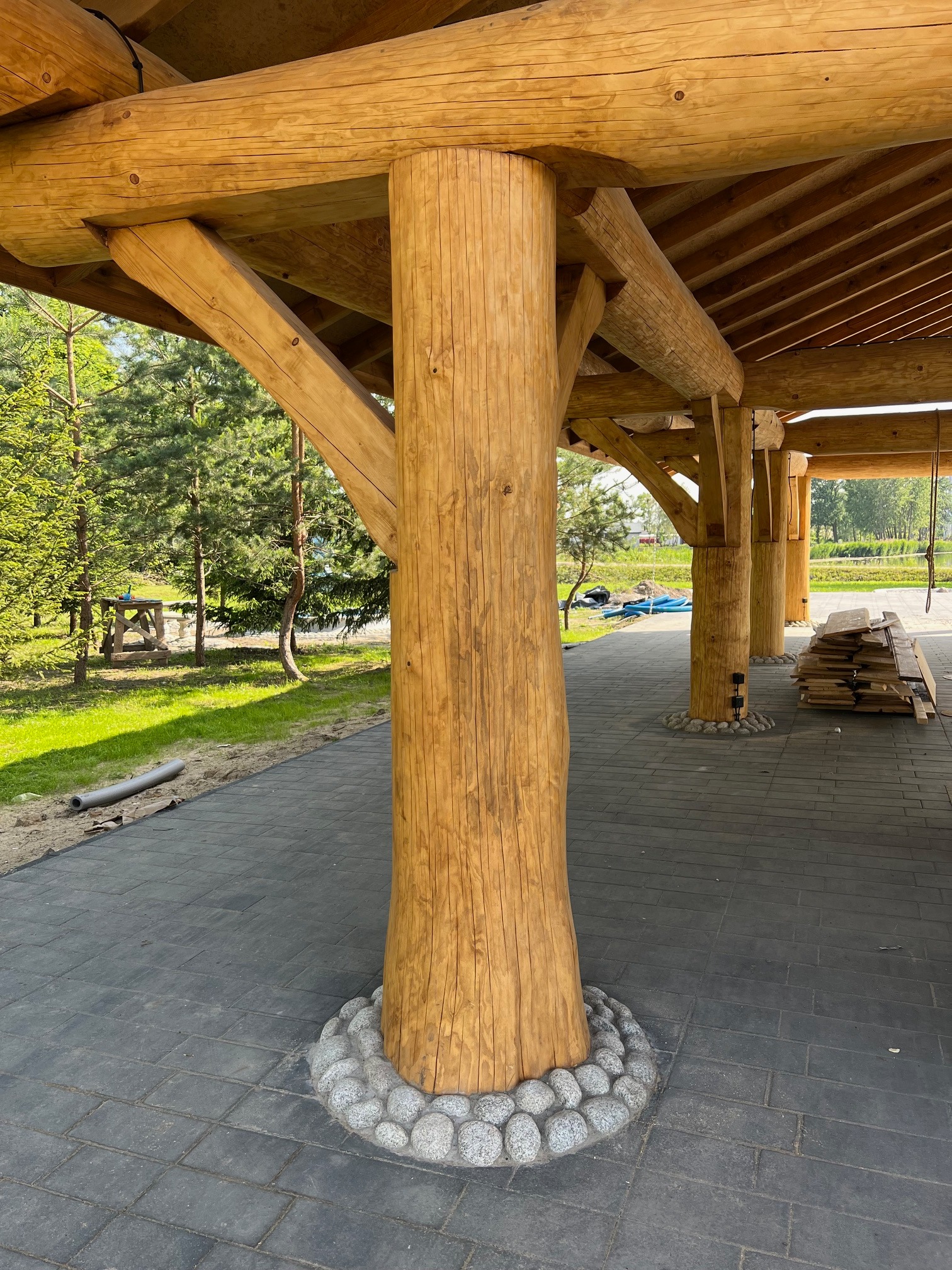 Drewniana kolumna podtrzymująca konstrukcję z bali, otoczona ozdobnymi kamieniami, na tle brukowanej nawierzchni i zieleni.