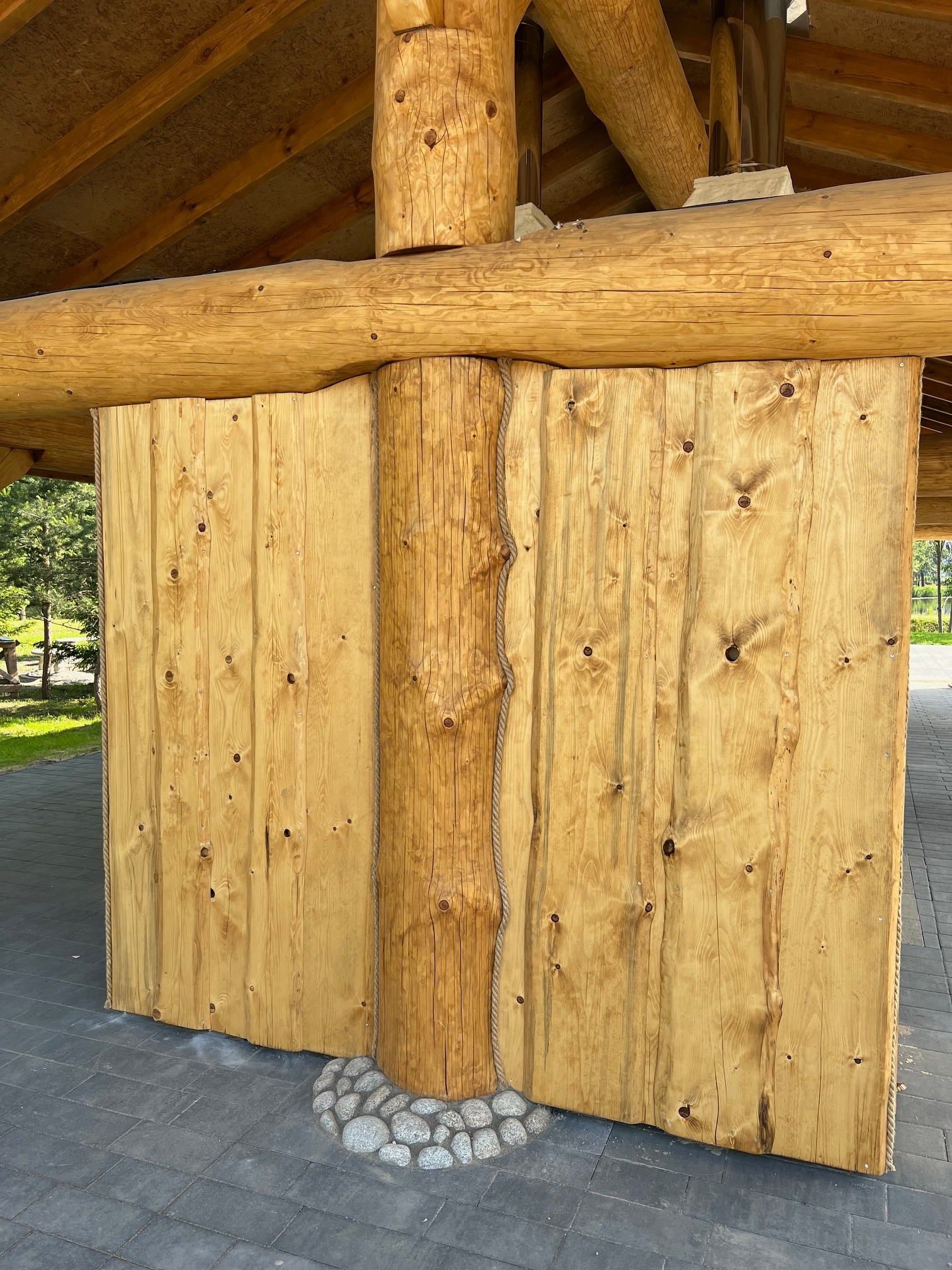 Drewniana konstrukcja altany z bali, widoczne połączenia bali i desek, podstawa kolumny z kamieni.