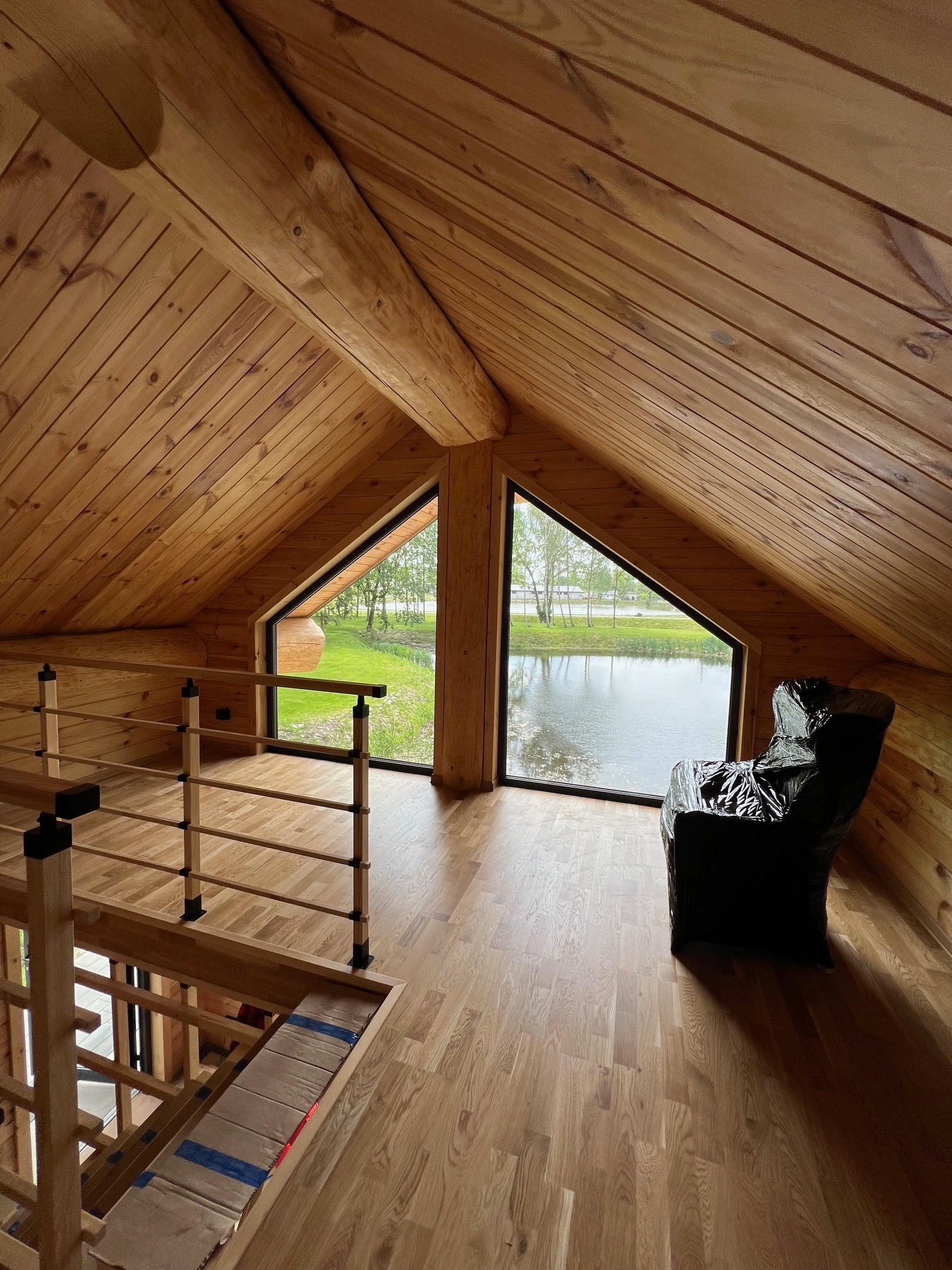 Wnętrze drewnianego domu z widokiem na jezioro przez trójkątne okno; krzesło zabezpieczone folią ochronną, drewniane schody z balustradą.