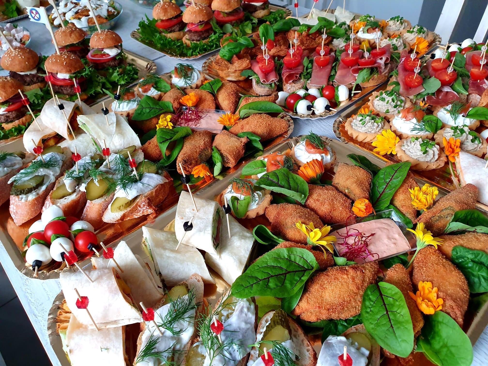 Bogato zastawiony stół z różnorodnymi przekąskami cateringowymi na złotych tacach: mini burgery z flagami, kanapeczki z wędliną i pomidorami, koreczki z mozzarelli i pomidorków koktajlowych...