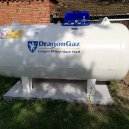 Biały zbiornik gazowy DragonGaz na betonowej podstawie, zainstalowany w ogrodzie we Wrocławiu, z widocznymi elementami przyłączeniowymi i tabliczką informacyjną.