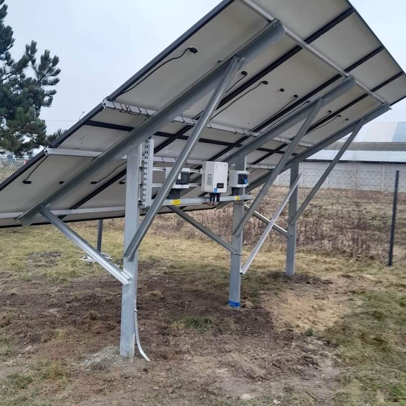Montaż paneli słonecznych: widok od spodu na stalową konstrukcję wsporczą z zamontowanymi mikroinwerterami, na tle zaoranej ziemi i ogrodzenia.