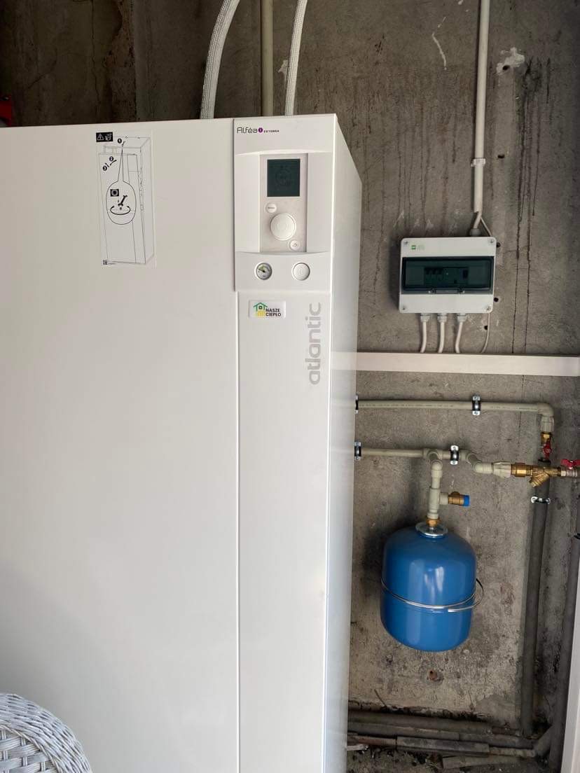 Biała pompa ciepła Atlantic Alfea Extensa z niebieskim zbiornikiem wyrównawczym, zamontowana na surowej, betonowej ścianie wewnątrz budynku.