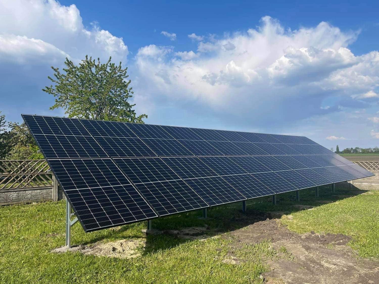 Panel słoneczny na metalowych wspornikach, zainstalowany na trawniku z widocznym ogrodzeniem i błękitnym niebem z chmurami w tle.