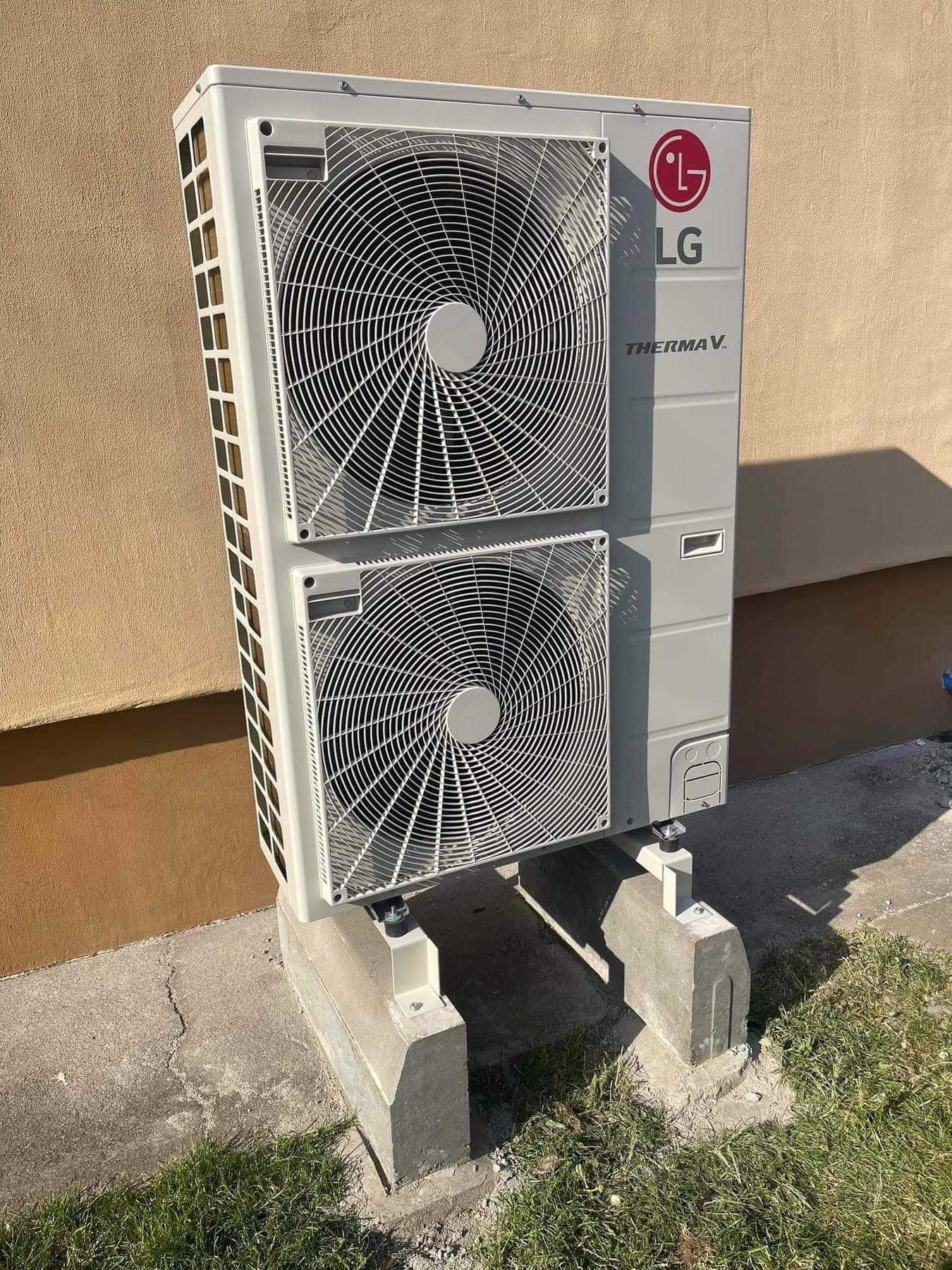 Zewnętrzna jednostka pompy ciepła LG Therma V, zamontowana na betonowych podstawach przy ścianie budynku, widoczne dwa wentylatory i logo producenta.