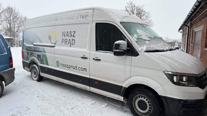 Biały samochód dostawczy firmy 'Nasz Prąd' zaparkowany na śniegu, z widocznymi płatkami śniegu na karoserii i dachu.