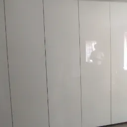 Biała, błyszcząca szafa wnękowa z widocznym odbiciem okna i osoby, zamontowana w surowym stanie pomieszczenia.