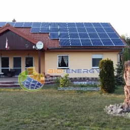 Dom jednorodzinny z panelami słonecznymi na dachu, polska flaga powiewająca na werandzie i drewniana rzeźba w ogrodzie. Widoczny logotyp firmy na trawniku.