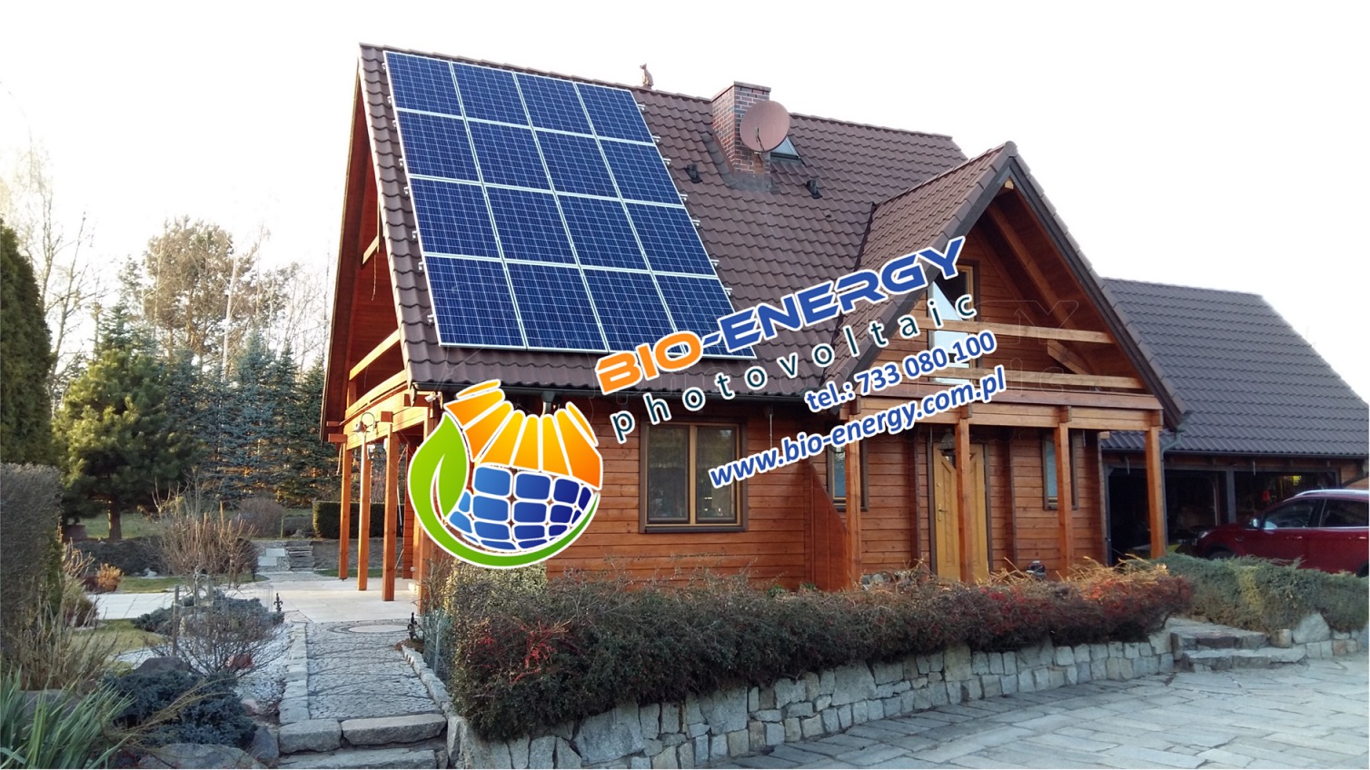 Drewniany dom jednorodzinny z panelami słonecznymi na dachu, widoczne logo firmy BIO-ENERGY photovoltaics na fasadzie.