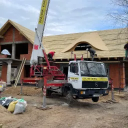 Budowa dachu z nietypowym oknem dachowym w kształcie łuku, ekipa dekarzy pracuje na rusztowaniu i podnośniku samochodowym, widoczny surowy stan budynku z czerwonej cegły i materiały budowlane...