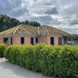 Budowa domu jednorodzinnego w trakcie: widoczna murowana konstrukcja z czerwonej cegły oraz drewniana więźba dachowa na tle pochmurnego nieba i zieleni drzew.