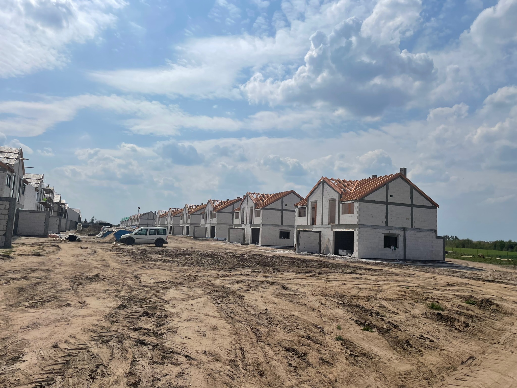 Szereg surowych, jednorodzinnych domów w trakcie budowy, z widoczną konstrukcją dachu pokrytą czerwoną dachówką, stojących wzdłuż piaszczystej drogi z wyraźnymi śladami opon, pod błękitnym niebem...