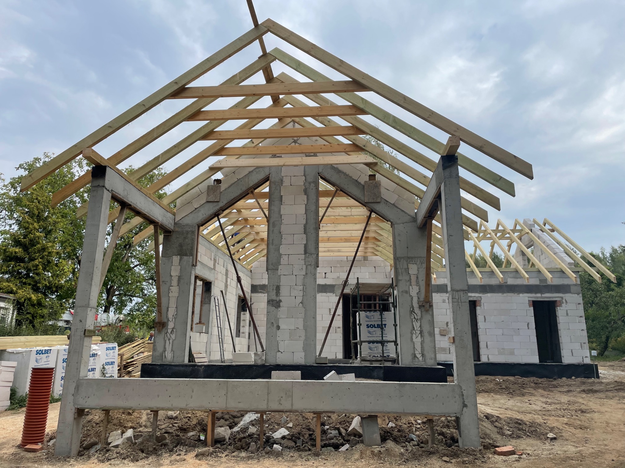 Surowy stan budowy domu jednorodzinnego z widoczną konstrukcją drewnianą dachu i betonowymi słupami nośnymi, materiały budowlane marki Solbet składowane obok.