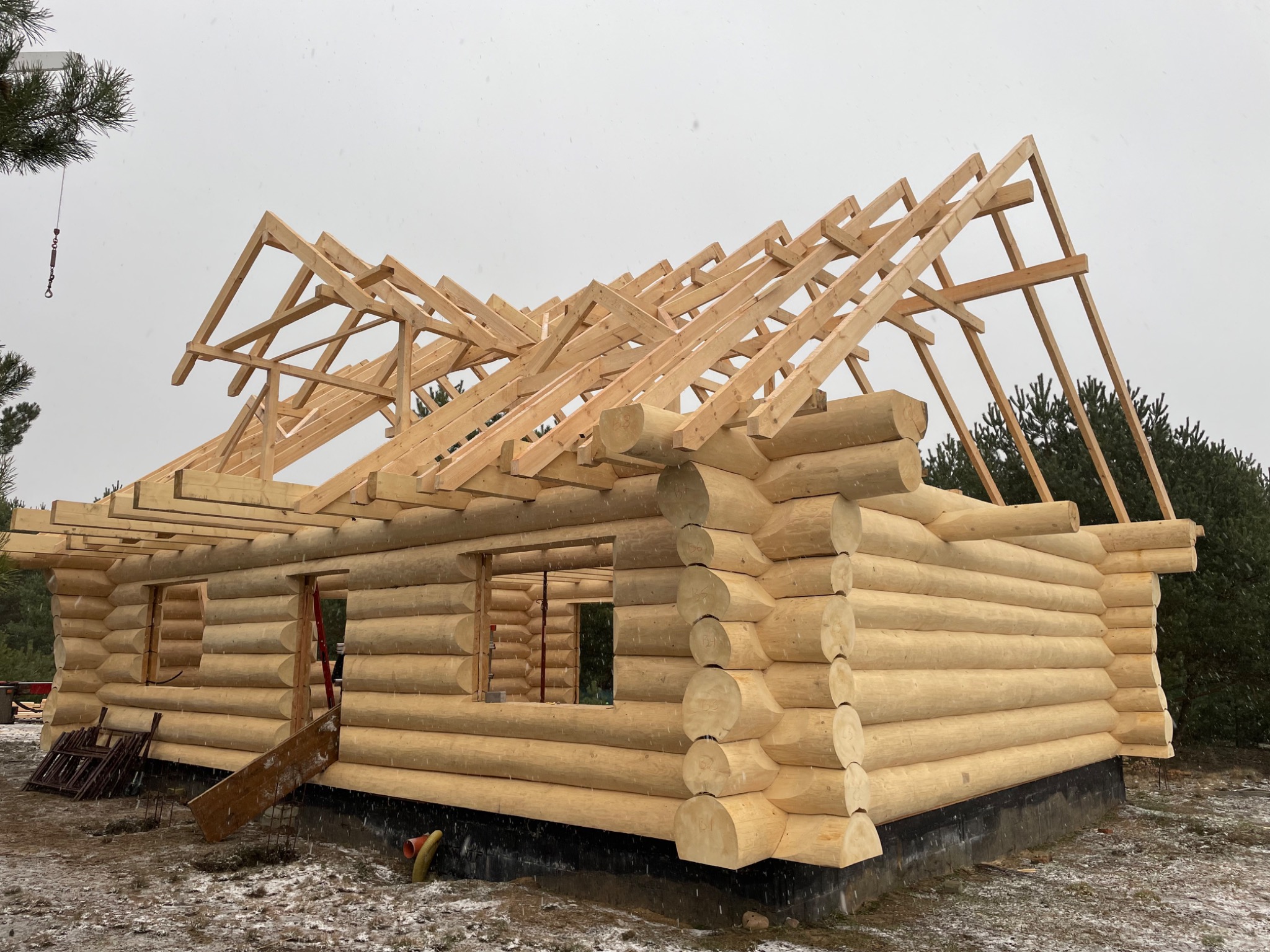 Surowa konstrukcja domu z bali w trakcie budowy, widoczna drewniana więźba dachowa na tle pochmurnego nieba.