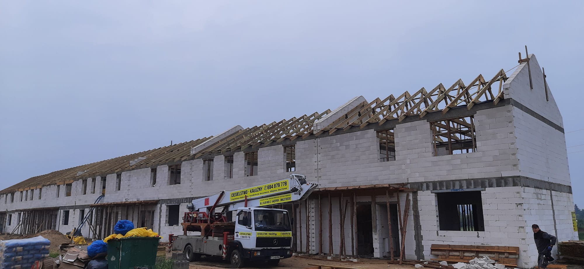 Budowa szeregu domów w stanie surowym otwartym, widoczna więźba dachowa i podnośnik koszowy z reklamą ciesielstwa.