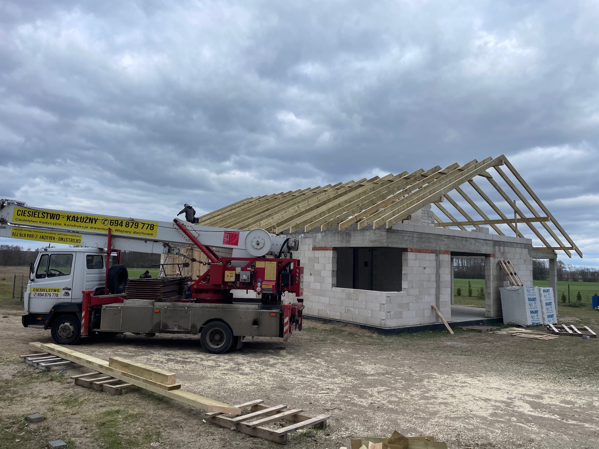 Budowa domu jednorodzinnego w stanie surowym otwartym, widoczna więźba dachowa, na pierwszym planie podnośnik z logo firmy ciesielskiej, palety z bloczkami SOLBET.