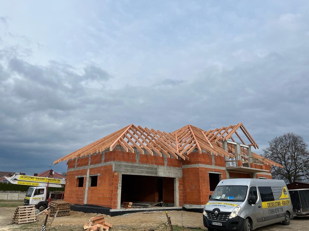 Budowa domu jednorodzinnego w trakcie realizacji, widoczna konstrukcja drewniana dachu, ściany z czerwonej cegły, na pierwszym planie zaparkowany bus firmy budowlanej i samochód dostawczy...