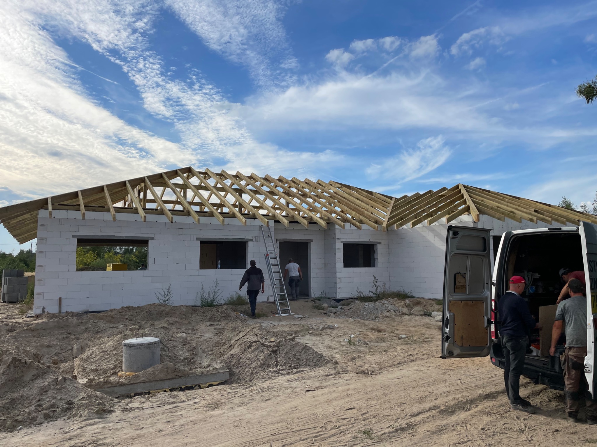 Budowa domu jednorodzinnego w trakcie montażu drewnianej konstrukcji dachu, widoczni pracownicy i samochód dostawczy na placu budowy, błękitne niebo z chmurami w tle.