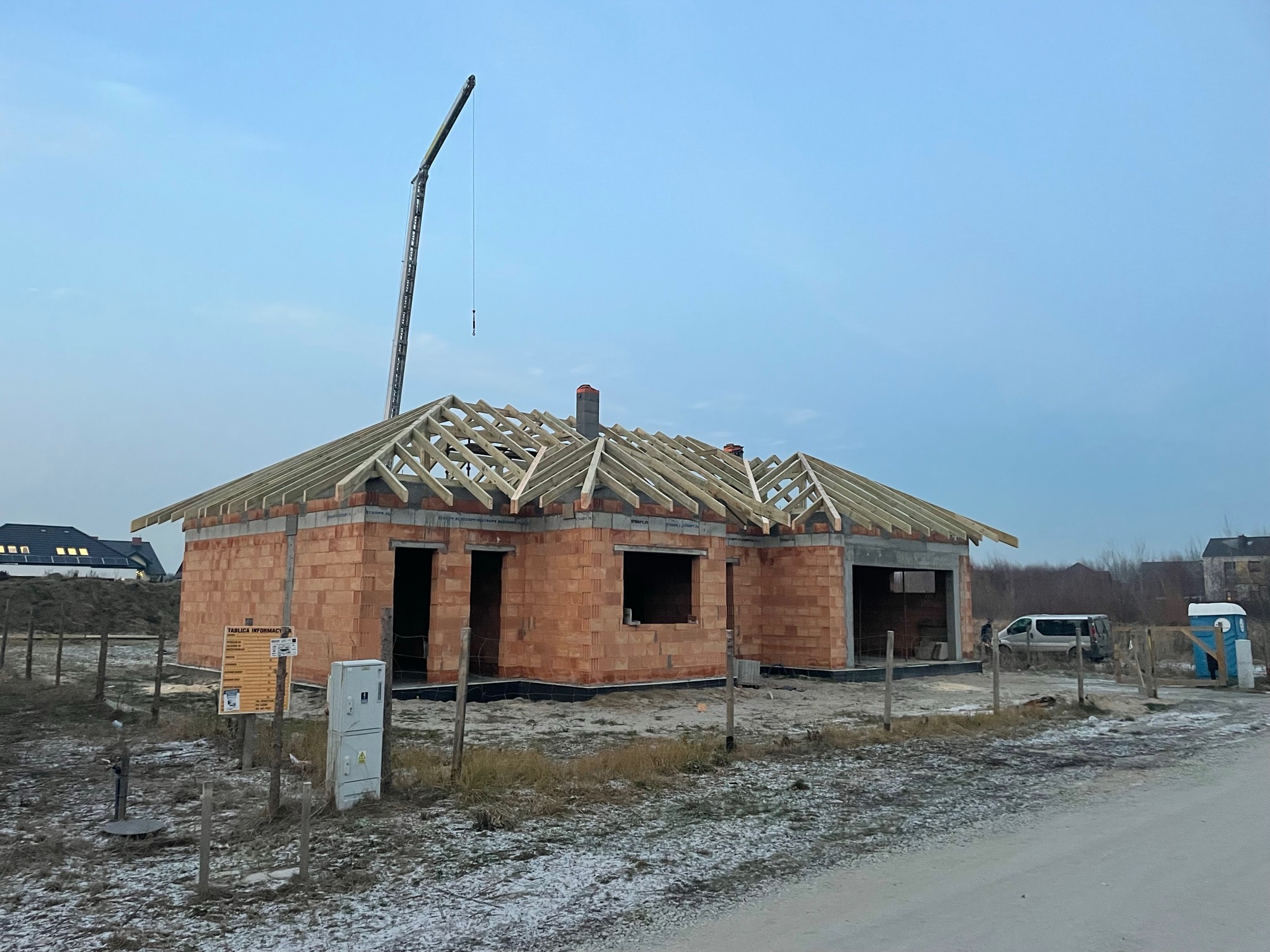 Budowa domu jednorodzinnego w stanie surowym otwartym, widoczna murowana konstrukcja ścian i drewniana więźba dachowa, z dźwigiem budowlanym w tle.