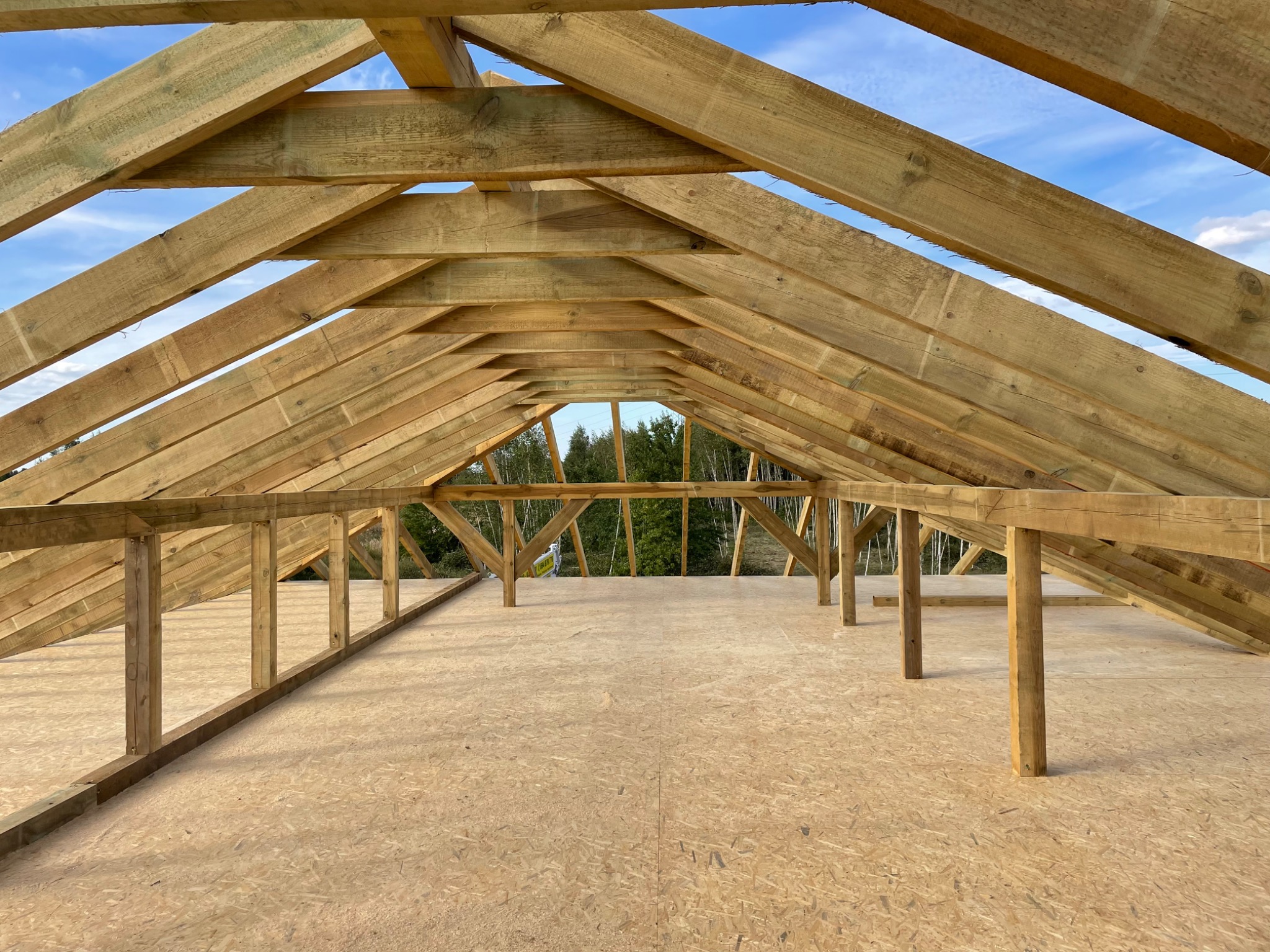 Konstrukcja drewniana dachu w trakcie budowy, widok od wewnątrz, z widoczną więźbą dachową i poszyciem z płyt OSB, na tle błękitnego nieba i zieleni drzew.