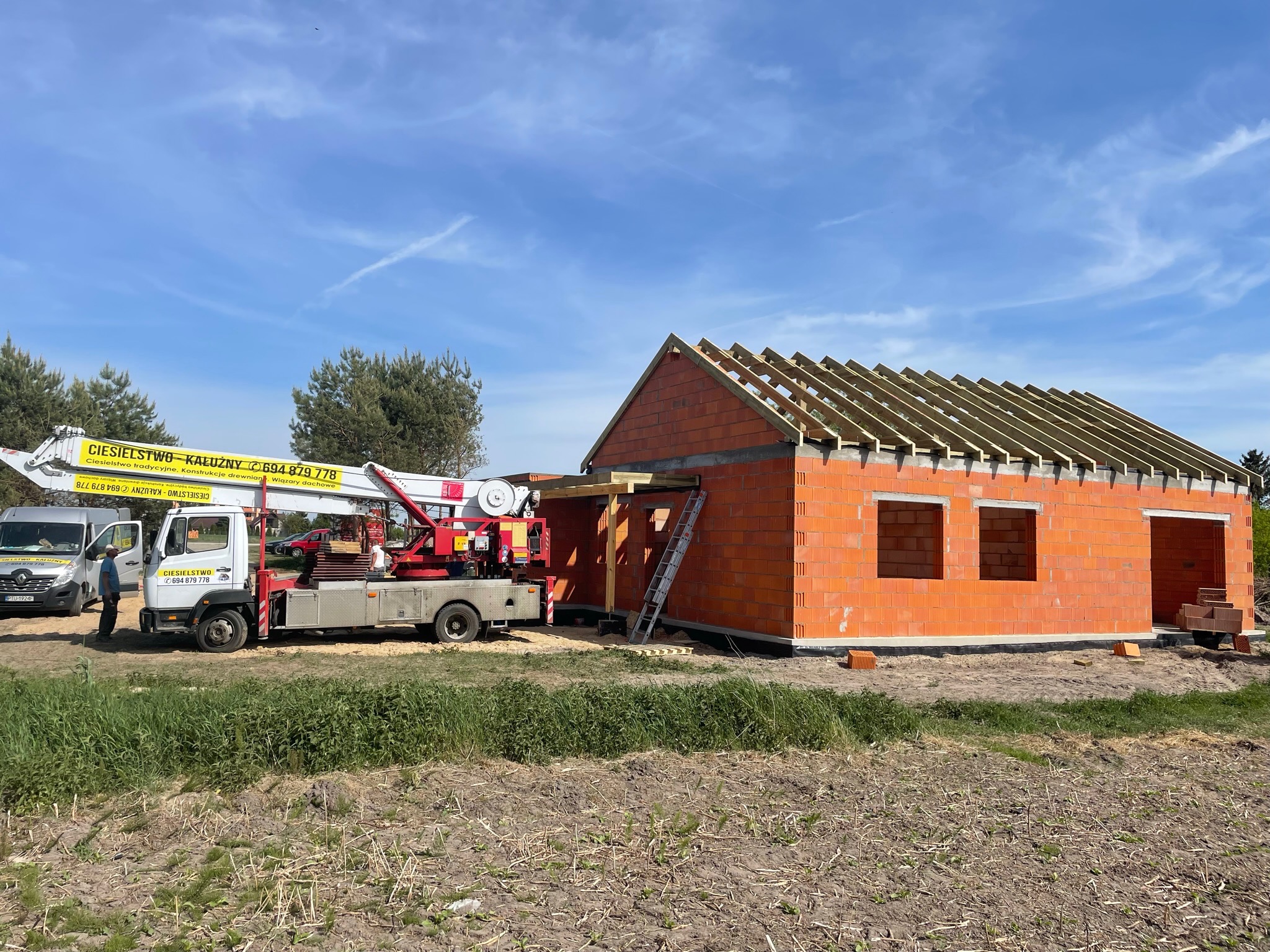 Wznoszony budynek z czerwonej cegły z częściowo wykonaną więźbą dachową, widoczny dźwig samochodowy z podnośnikiem firmy ciesielskiej i drabina oparta o ścianę budynku.