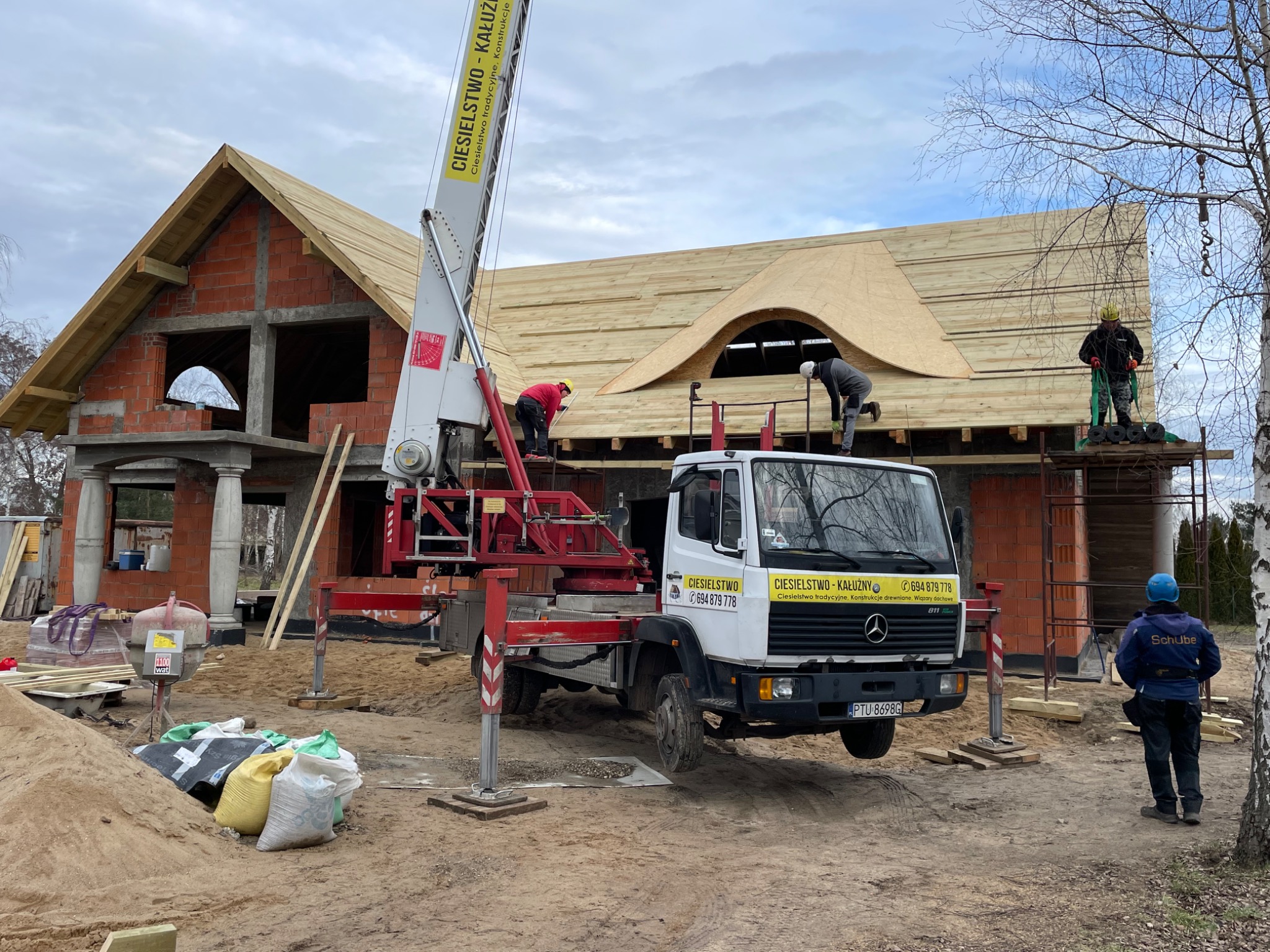 Budowa dachu z nietypowym oknem dachowym w kształcie łuku, ekipa dekarzy pracuje na rusztowaniu i podnośniku samochodowym, widoczny surowy stan budynku z czerwonej cegły i materiały budowlane...