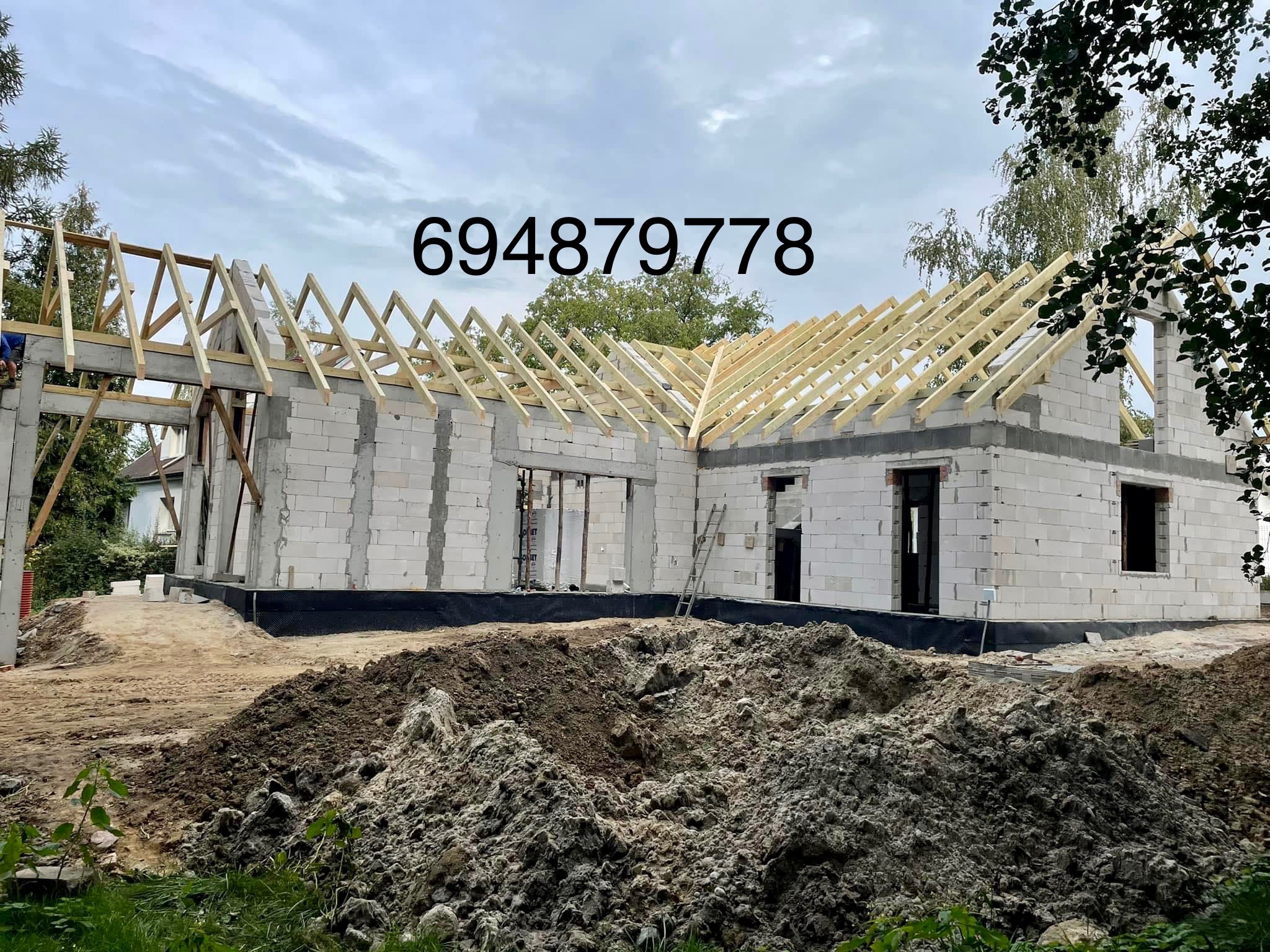 Wznoszenie drewnianej konstrukcji dachu na surowym budynku z białych bloczków, widoczne elementy deskowania i ziemia wokół budynku.