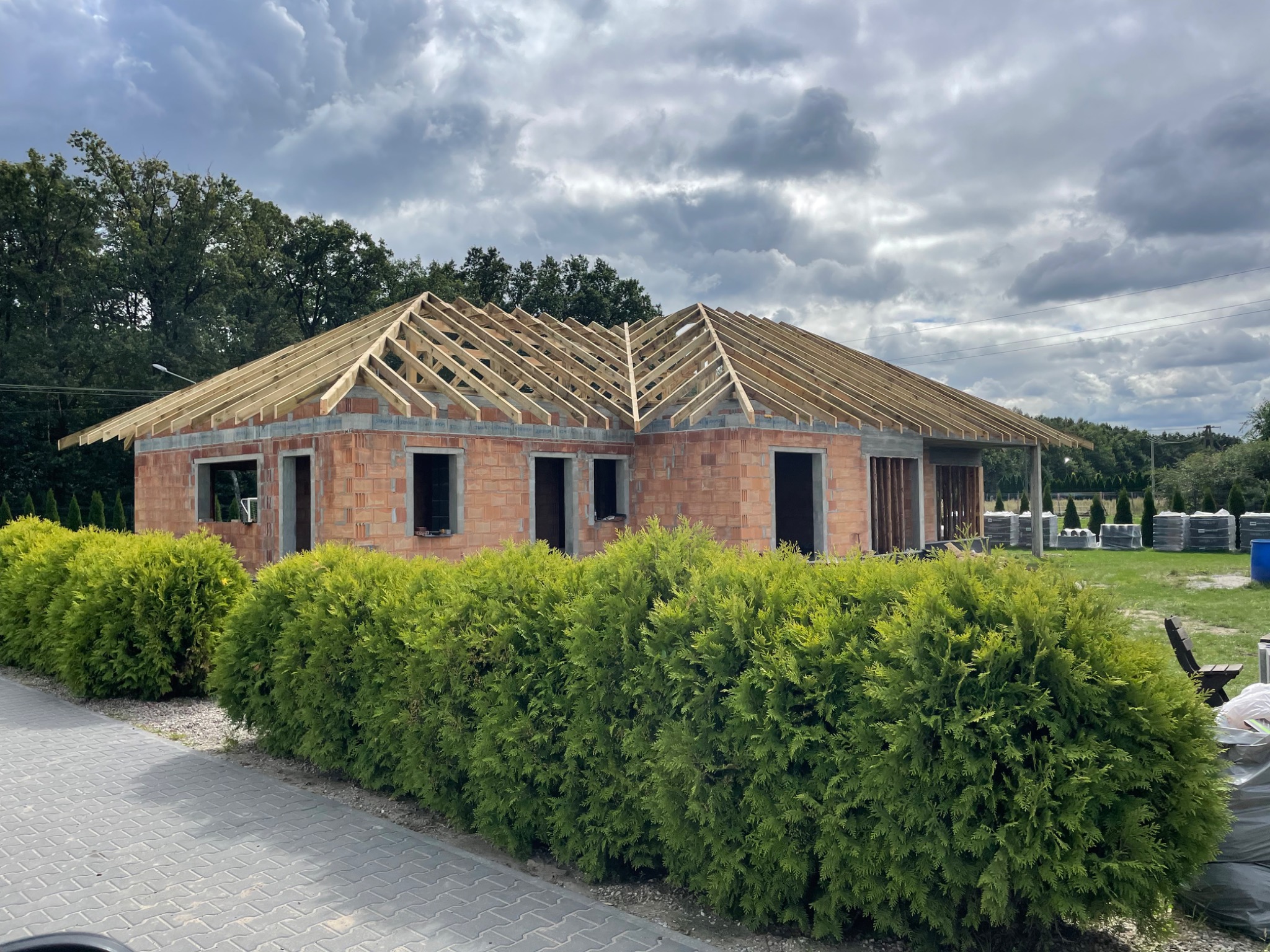 Budowa domu jednorodzinnego w trakcie: widoczna murowana konstrukcja z czerwonej cegły oraz drewniana więźba dachowa na tle pochmurnego nieba i zieleni drzew.