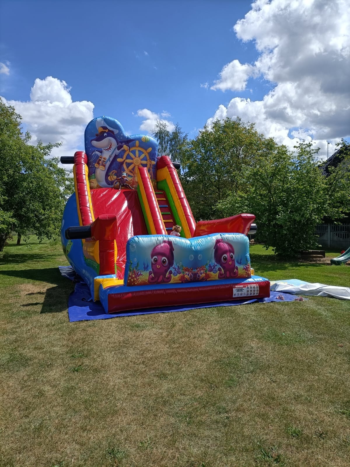 Kolorowa, dmuchana zjeżdżalnia z motywami morskimi (rekin-kapitan, ośmiornice) ustawiona na trawniku w ogrodzie.