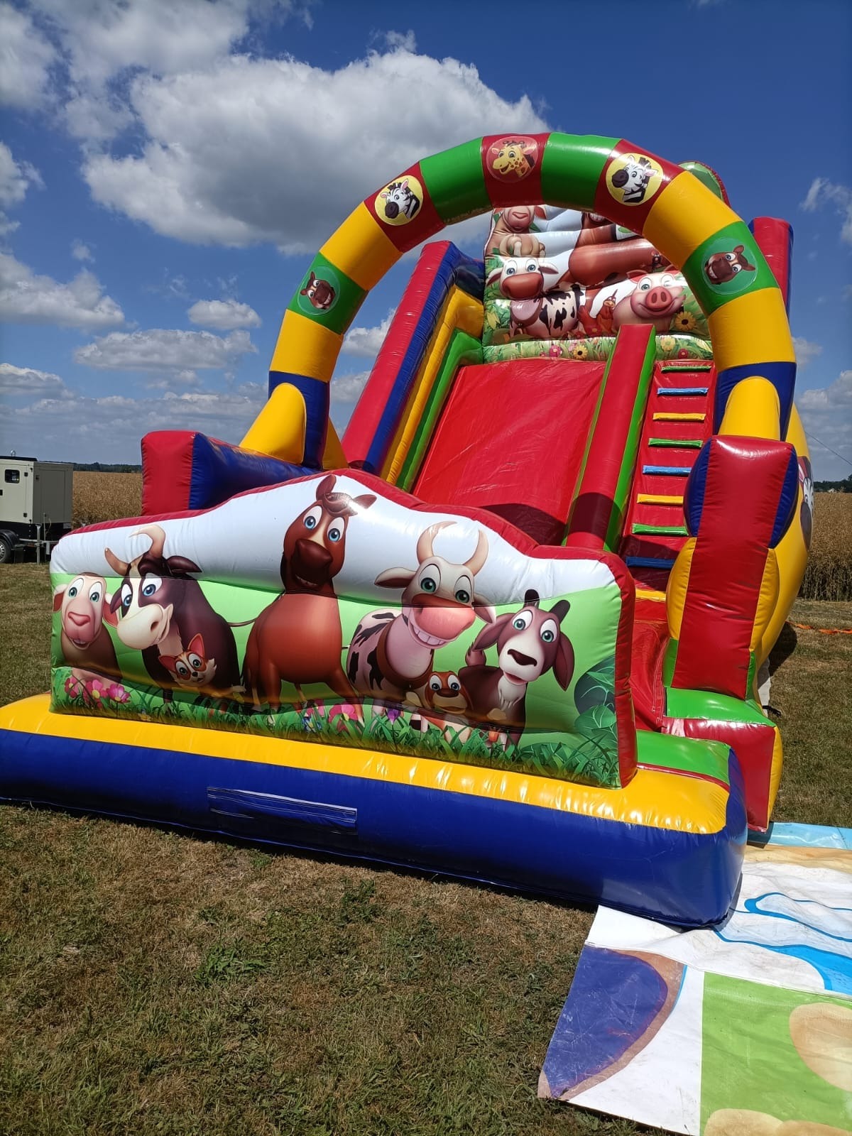 Kolorowa, nadmuchiwana zjeżdżalnia z motywem zwierząt gospodarskich na trawie, z widocznymi elementami konstrukcyjnymi i dekoracyjnymi.
