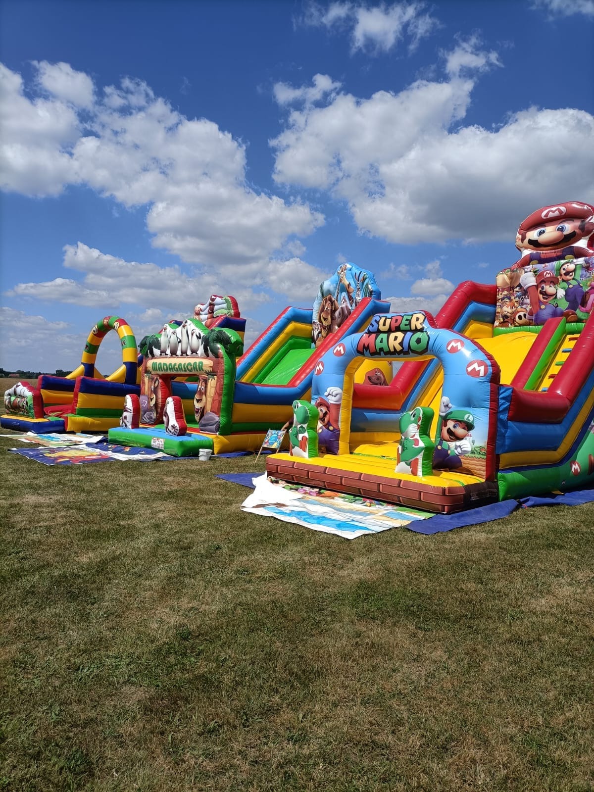 Dwa kolorowe, dmuchane place zabaw na trawie, jeden z motywem Madagaskaru, drugi z motywem Super Mario, pod błękitnym niebem z białymi chmurami.