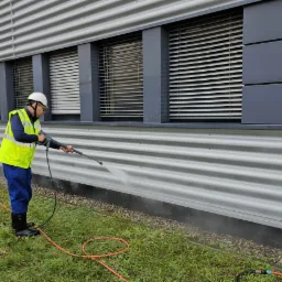 Pracownik w kasku i kamizelce odblaskowej myje elewację przemysłową myjką ciśnieniową, widoczne zacieki i trawa.