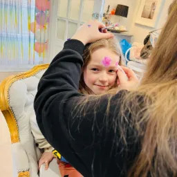 Dziewczynka siedzi na fotelu podczas malowania różowego kwiatka na jej czole pędzlem. W tle dekoracje urodzinowe.