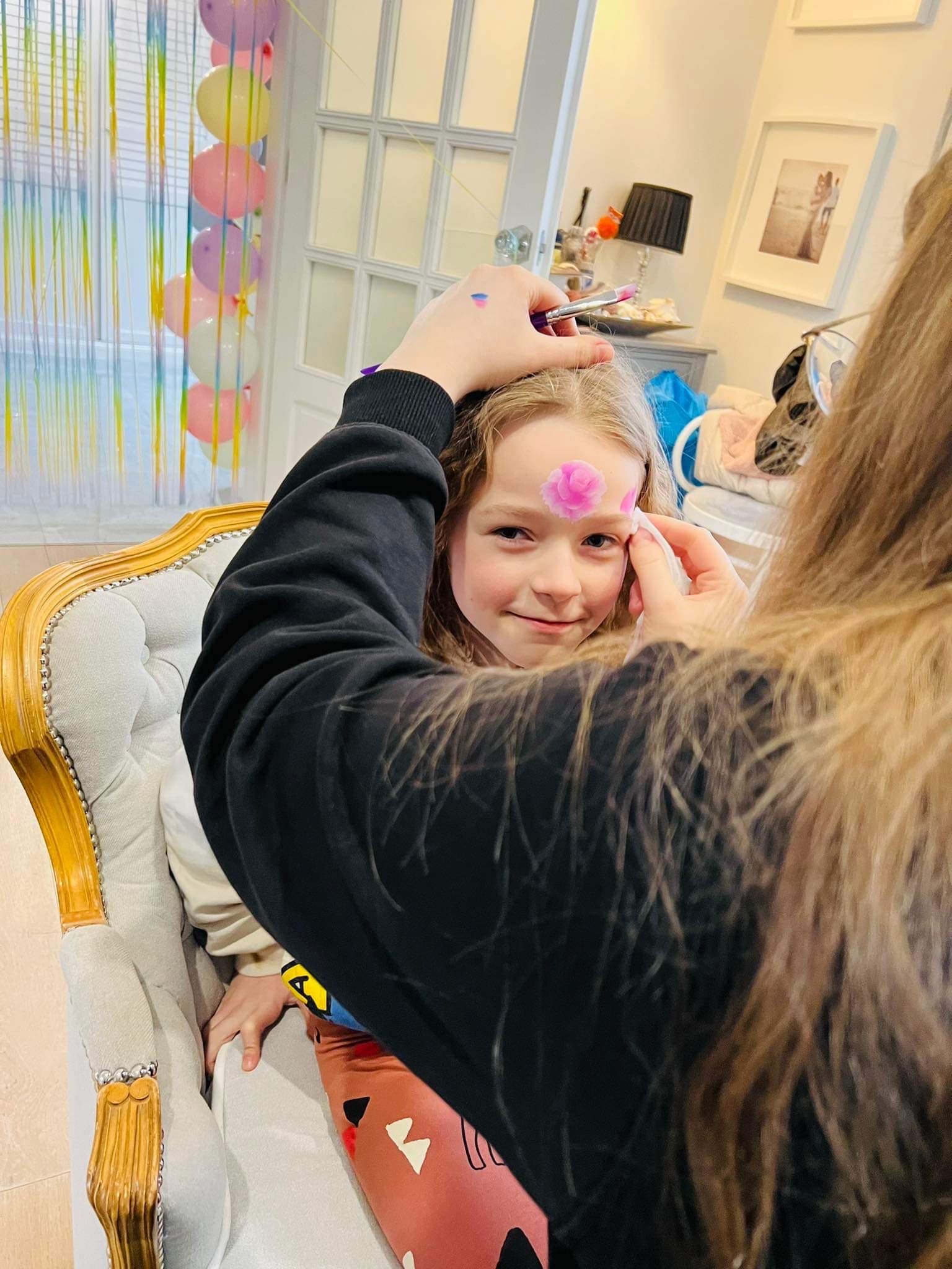 Dziewczynka siedzi na fotelu podczas malowania różowego kwiatka na jej czole pędzlem. W tle dekoracje urodzinowe.