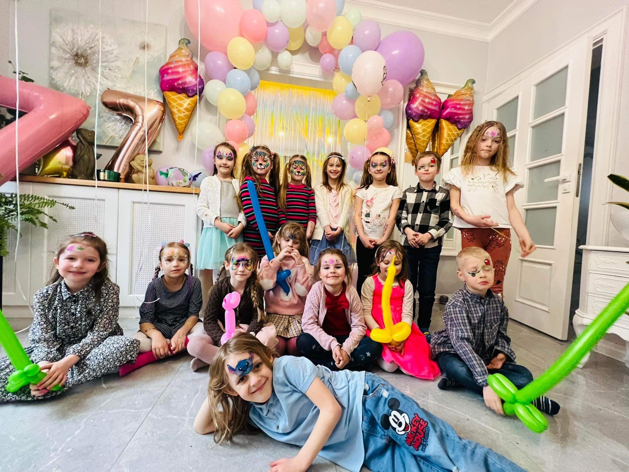 Grupa dzieci z pomalowanymi twarzami pozuje na tle dekoracji balonowych w kształcie lodów i cyfr na przyjęciu urodzinowym.