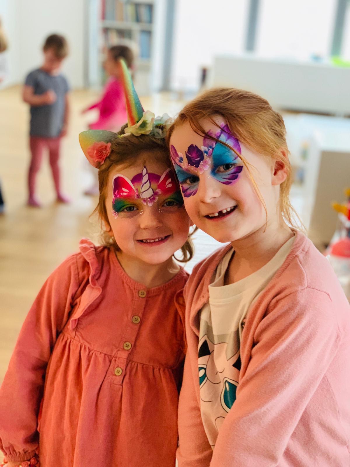 Dwie uśmiechnięte dziewczynki z pomalowanymi twarzami: jedna z motywem jednorożca z tęczową grzywą i brokatem, druga z niebiesko-fioletowym motywem kwiatowym wokół oczu, w tle dzieci i regał...