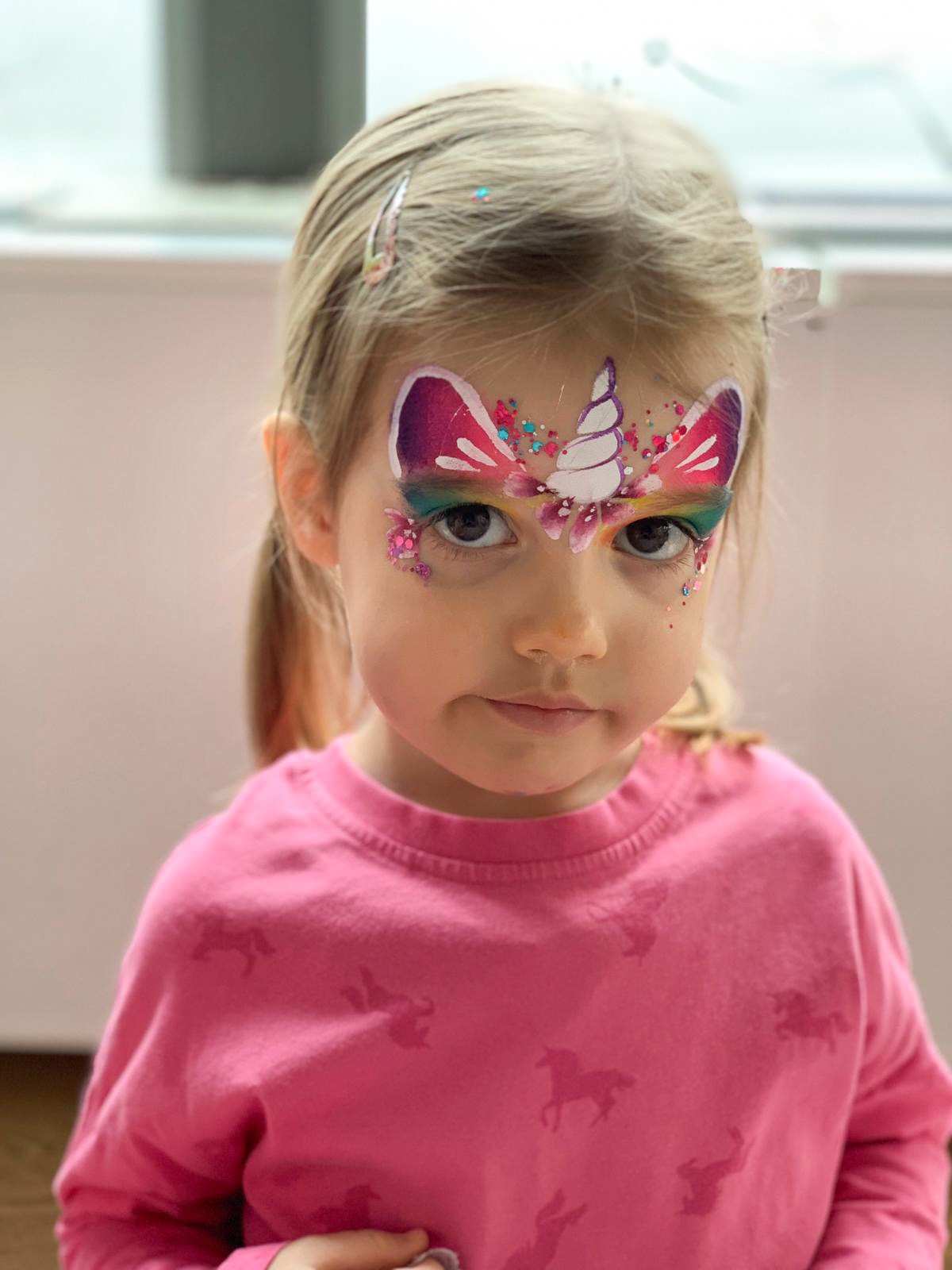 Dziewczynka z pomalowaną twarzą w motywie jednorożca, z brokatem i kolorowymi cieniami wokół oczu, ubrana w różową bluzkę z nadrukiem jednorożców.