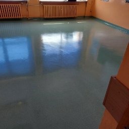 Błyszcząca, świeżo umyta podłoga z linoleum w pustym pomieszczeniu z pomarańczowymi ścianami i widocznymi grzejnikami pod oknami.