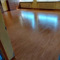 Wypolerowana, pusta sala z drewnianą podłogą, brązowymi szafkami przy ścianie i dwoma grzejnikami pod oknami. Podłoga odbija światło, tworząc jasne refleksy.