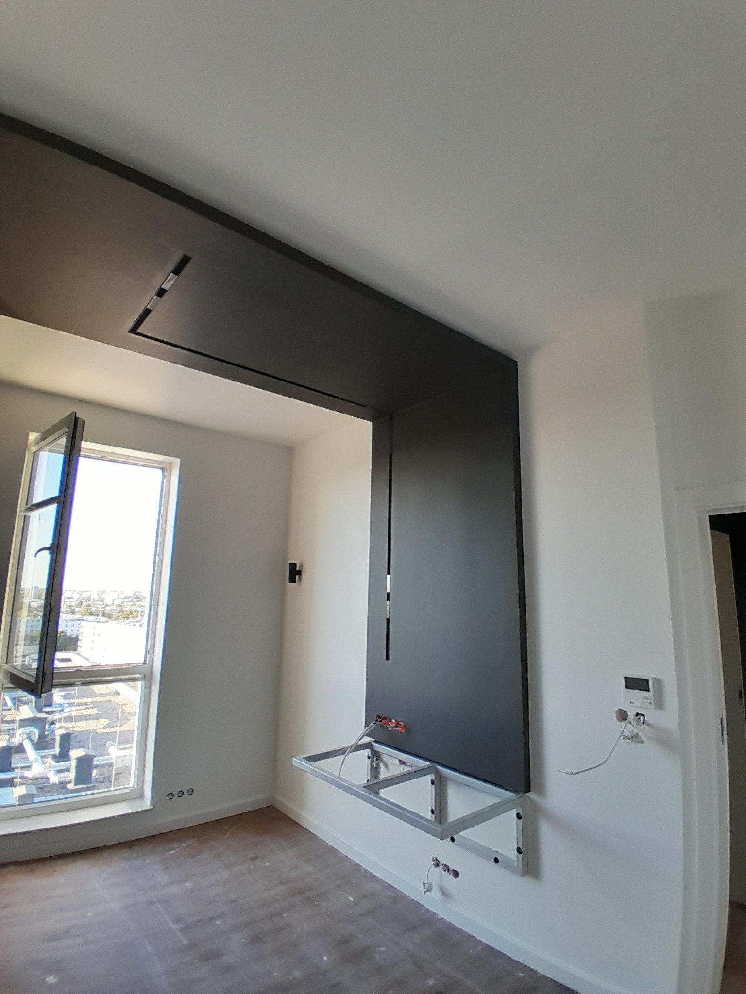 Wnętrze pokoju z szarą zabudową meblową na ścianie, widoczne przygotowania do montażu lub szpachlowania, aluminiowy stelaż pod zabudową, otwarte okno z widokiem na miasto.