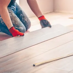 Pracownik w czerwonych rękawicach montuje panel podłogowy na różowym podkładzie, obok leży żółta miarka.