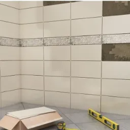 Proces układania beżowych płytek ściennych w rogu pomieszczenia, z dekoracyjnym pasem płytek z motywem roślinnym, widoczne narzędzia: poziomica i miarka.